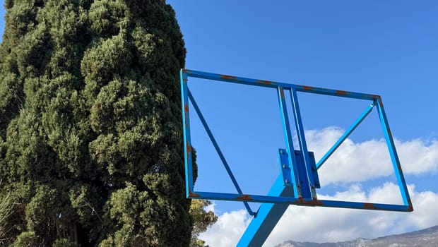 Rusty, hollow blue basketball hoop frame against tall evergreen tree and blue sky. High quality photo