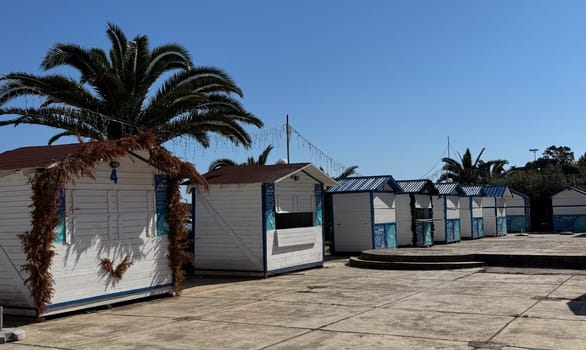 Budva,Montenegro-April 11,2025:Row of small, closed white wooden beach market huts with palm trees and string lights on a sunny day.