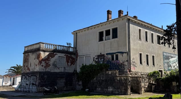 Budva,Montenegro-April 11,2025:Old two-story building with peeling paint, graffiti, and boarded windows under clear sky, surrounded by palm trees.