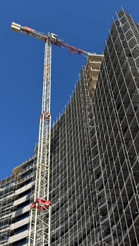 Budva, Montenegro - April 11,2025:Tower crane beside a high-rise under construction, fully wrapped in scaffolding against a deep blue sky.