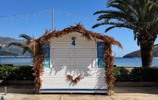 Budva,Montenegro-April 11,2025A small white fair house number 4, decorated with dried plants, stands against a picturesque beach and island backdrop.