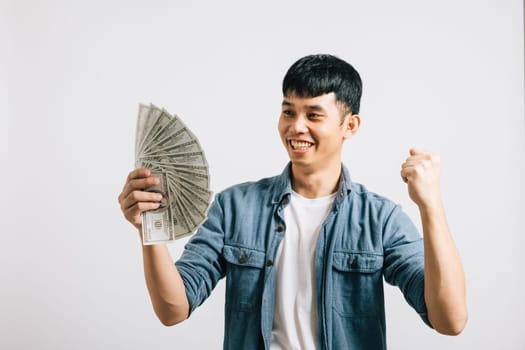Portrait smiling Asian young man holding a bunch of money and pointing studio shot isolated white background, happiness and excitement man in dollar banknotes celebrating his financial success