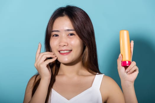 Beautiful Asian young woman holding body lotion on hand in studio shot isolated on blue background, Smiling brunette female hold cream sunblock bottle, tube of sunscreen, protect sun light and UV