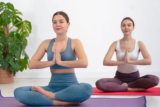 Caucasian women practicing yoga in seated lotus pose with palms together, wearing sportswear on mats in a bright indoor space. Calm expression and focus reflect mindfulness and healthy lifestyle