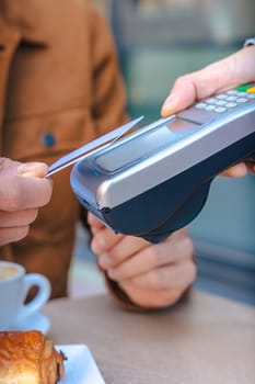 Close-up of two people making a contactless card payment at a cafe table with a POS terminal. Coffee and pastry are visible. Modern lifestyle and digital finance concept in urban setting. Vertical
