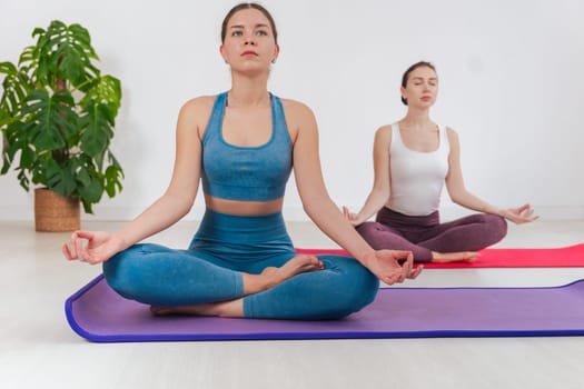 Two caucasian women doing yoga meditation indoors, sitting in lotus pose on mats. Calm and focused atmosphere in a bright minimal room, promoting mindfulness and physical wellness.