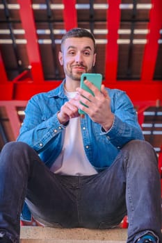 Happy man in denim jacket reacts with excitement while looking at his smartphone. Bright red architectural background in urban setting. Candid expression and joyful moment captured outdoors. Vertical