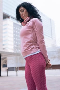 Caucasian adult woman with curly black hair stretching her arms outdoors in an urban setting.she is wearing a pink sweatshirt and exercising for a healthy lifestyle, fitness, and wellness in the city