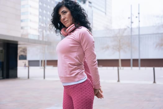 Caucasian adult woman with curly black hair stretching her arms outdoors in an urban setting.she is wearing a pink sweatshirt and exercising for a healthy lifestyle, fitness, and wellness in the city