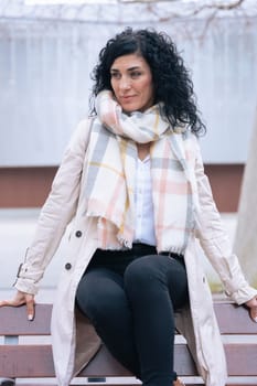 Vertical portrait of a woman sitting on a bench outdoors. She wears a beige coat and scarf, looking calm and confident. Urban background with winter atmosphere and seasonal fashion concept. Vertical