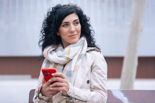 Happy caucasian woman standing outdoors in urban setting, wearing coat and scarf, using her red smartphone. She is smiling and relaxed, enjoying a casual moment on a cold day in the city.