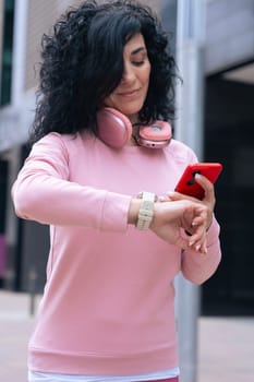 Middle adult Caucasian woman with curly black hair checking her smartwatch outdoors. She is wearing a pink sweatshirt and pink headphones, representing fitness, technology, and an active lifestyle.