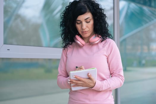 Caucasian sporty woman in a pink hoodie writing in a notebook outdoors. Wearing headphones around her neck, she appears focused and relaxed after a workout in an urban setting.