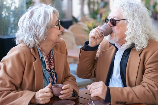 Caucasian senior couple enjoying coffee at an outdoor cafe. The woman speaks closely to the man, creating an intimate and thoughtful moment. Concept of connection, trust, and shared experiences.