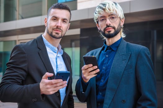 Portrait of two confident businessmen serious and holding smartphones, wearing formal suits, standing outdoors near modern corporate office buildings, looking at camera