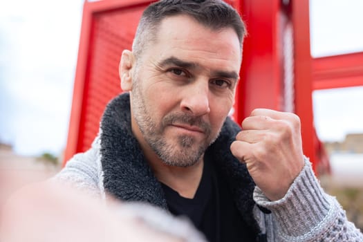 Confident caucasian man looking at camera with a raised fist in front of a red structure. Strong expression and casual winter outfit in an urban environment convey power, determination and energy