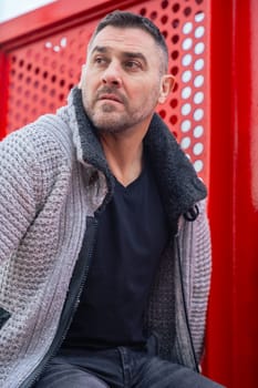 Portrait of a serious caucasian man standing by a bold red metal structure in an urban environment. Wearing a gray sweater and black shirt, he looks confidently at the camera with city view behind