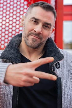 Caucasian man smiling slightly and making a peace sign with his hand. Wearing casual clothes in front of a red perforated urban structure, expressing confidence, calmness, and street style vibe