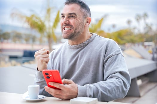 Excited caucasian man reacts with joy after winning an online game bet on his smartphone, sitting at an outdoor cafe table with coffee, enjoying his moment of triumph and happiness