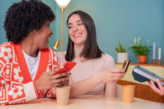 African American and Caucasian women smiling while paying at a coffee shop. One holds a card, the other a phone. Concept of friendship, technology, and casual lifestyle in a modern setting.