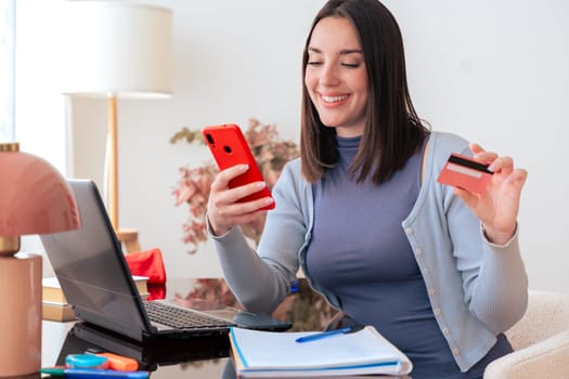 Happy young caucasian woman using smartphone and credit card for online shopping at home. She sits at a desk with a laptop and notebook, enjoying a seamless digital shopping experience.