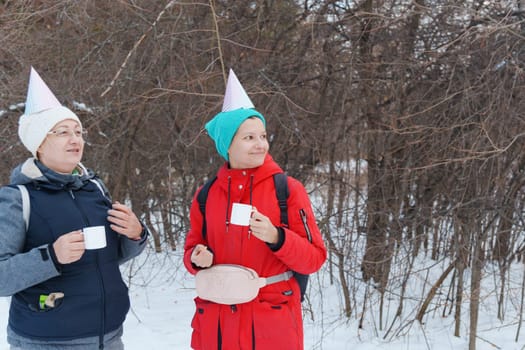 Two friends in festive party hats enjoy warm beverages while standing in a snowy area.