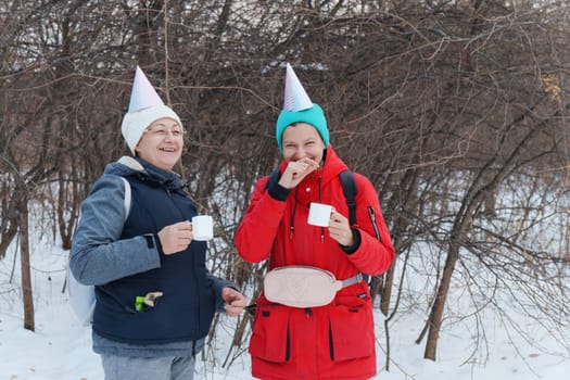 Two friends in festive party hats enjoy warm beverages while standing in a snowy area.