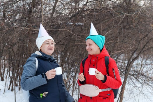 Two friends in festive party hats enjoy warm beverages while standing in a snowy area.