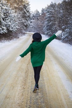 A young girl in a green sweater and hat walks in the middle of a snowy road in a thick pine forest. Freezing day