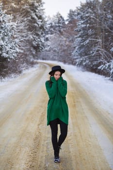 A young girl in a green sweater stands in the middle of a snowy road in a thick pine forest. Freezing day