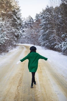 A young girl in a green sweater and hat walks in the middle of a snowy road in a thick pine forest. Freezing day
