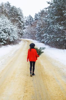 A girl in a red sweater and hat stands in the middle of a snowy road in a forest with pine branches. Weekend trip