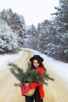 A girl in a red sweater and hat, in red mittens holds a basket with pine branches in her hands stands in the middle of a snow-covered road in a forest with pine branches