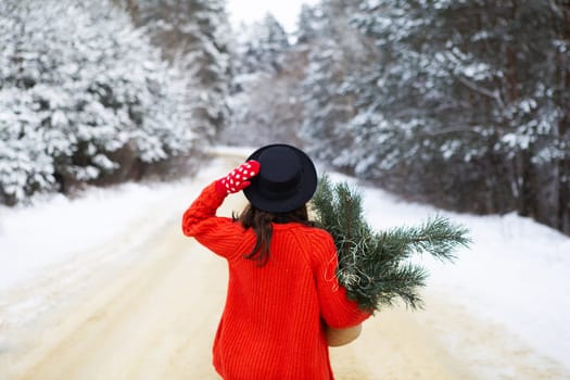 A girl in a red sweater and hat stands in the middle of a snowy road in a forest with pine branches. Weekend trip
