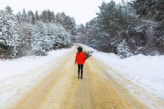 A girl in a red sweater and hat stands in the middle of a snowy road in a forest with pine branches. Weekend trip