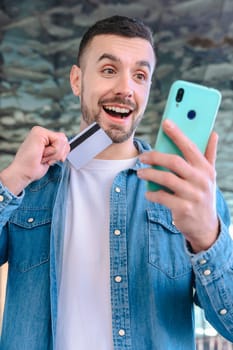 Happy young man using his smartphone while holding a credit card. He smiles with excitement, standing outside a glass building, enjoying an online shopping or payment experience. Vertical