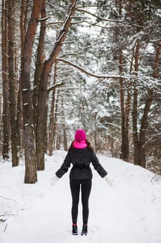 A girl in a pink scarf and hat stands with her back in the middle of a pine forest. Beautiful pine forest on a frosty day