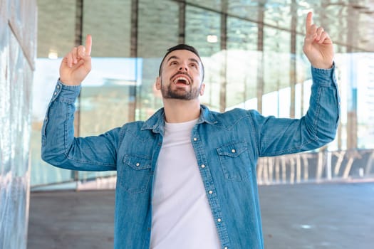 Smiling caucasian man in denim jacket points upward with both hands. He stands outside a modern glass building, expressing joy, excitement, and positivity in a stylish and urban environment.