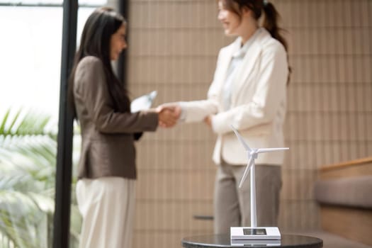 In a modern office setting, two businesswomen shake hands, symbolizing their commitment to sustainability while featuring a model of a wind turbine,