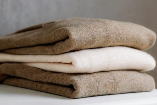 Three pieces of thin warm sweaters in nude shades made of natural wool lie flatly folded on a white shelf, side view hyper close-up. Concept women's clothing, new collection, natural things, fashion industry.