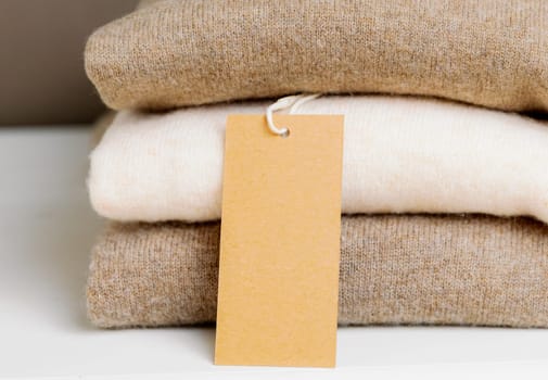 Three pieces of thin sweaters of nude shades made of natural wool with an empty kraft tag lie neatly folded in a stack on a white wooden shelf, side view very close up. Concept women's clothing, new collection, natural things, fashion industry.