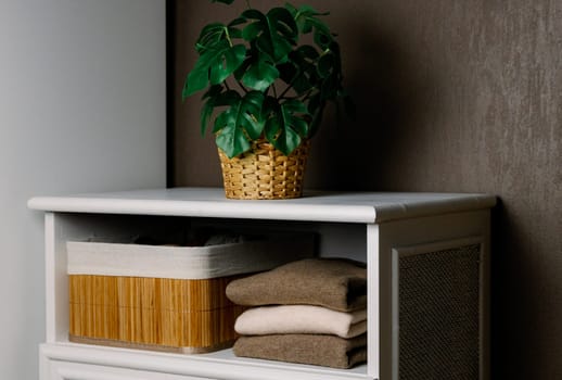 Thin fashionable sweaters in nude shades made from natural goat hair lie folded in a stack on a shelf of a retro white closet next to a bamboo basket and a flower in a pot against a brown wall, medium shot side view. Conceptual women's clothing, new collection, natural items, fashion industry.