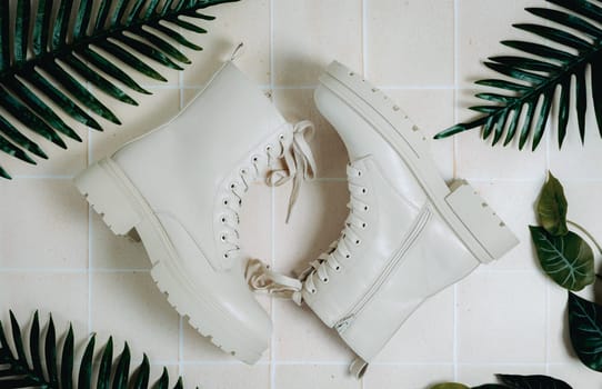 Beautiful view of a pair of fashionable stylish women's classic cream-colored boots with rough soles and lace-ups lie on a tiled floor with green palm leaves, flat lay close-up. Concept of women's shoes, fashion collection.