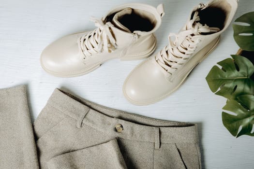 One pair of fashionable stylish women's classic cream-colored boots with rough soles and lace-ups with pants on a light cement floor with a monstera flower, close-up top view. Concept of women's shoes and clothing, fashion collection.