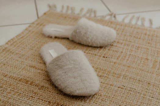 One pair of beautiful indoor light beige shaggy soft, fashionable and warm slippers stand on a natural jute rug on the tiled floor in the room, top side view close-up with depth of field. House shoes concept.
