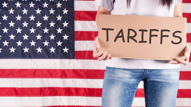 A young woman stands before an American flag, holding a sign that says tariffs in black marker. She wears a white shirt and blue jeans. The photo is from a low angle, concealing her face.