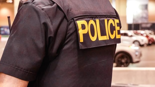 A police officer in uniform with a gun on his belt is standing with his back turned to the camera. The officer is wearing a black uniform with a badge on the shoulder.