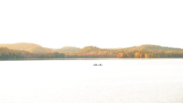 The image shows a small boat on a calm lake with a mountain range in the background. The boat is rowed by two people and the sun is rising or setting, casting a golden glow over the scene.