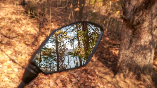 Looking through the rearview mirror of a motorcycle, you see a beautiful view of a lake surrounded by trees in the distance. The dirt road through the forest is covered with fallen leaves.