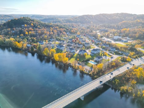 A beautiful small town filled with charming houses, vibrant trees, and a serene river flowing through it. The sky is a brilliant blue adorned with fluffy white clouds, and the sun shines brightly.